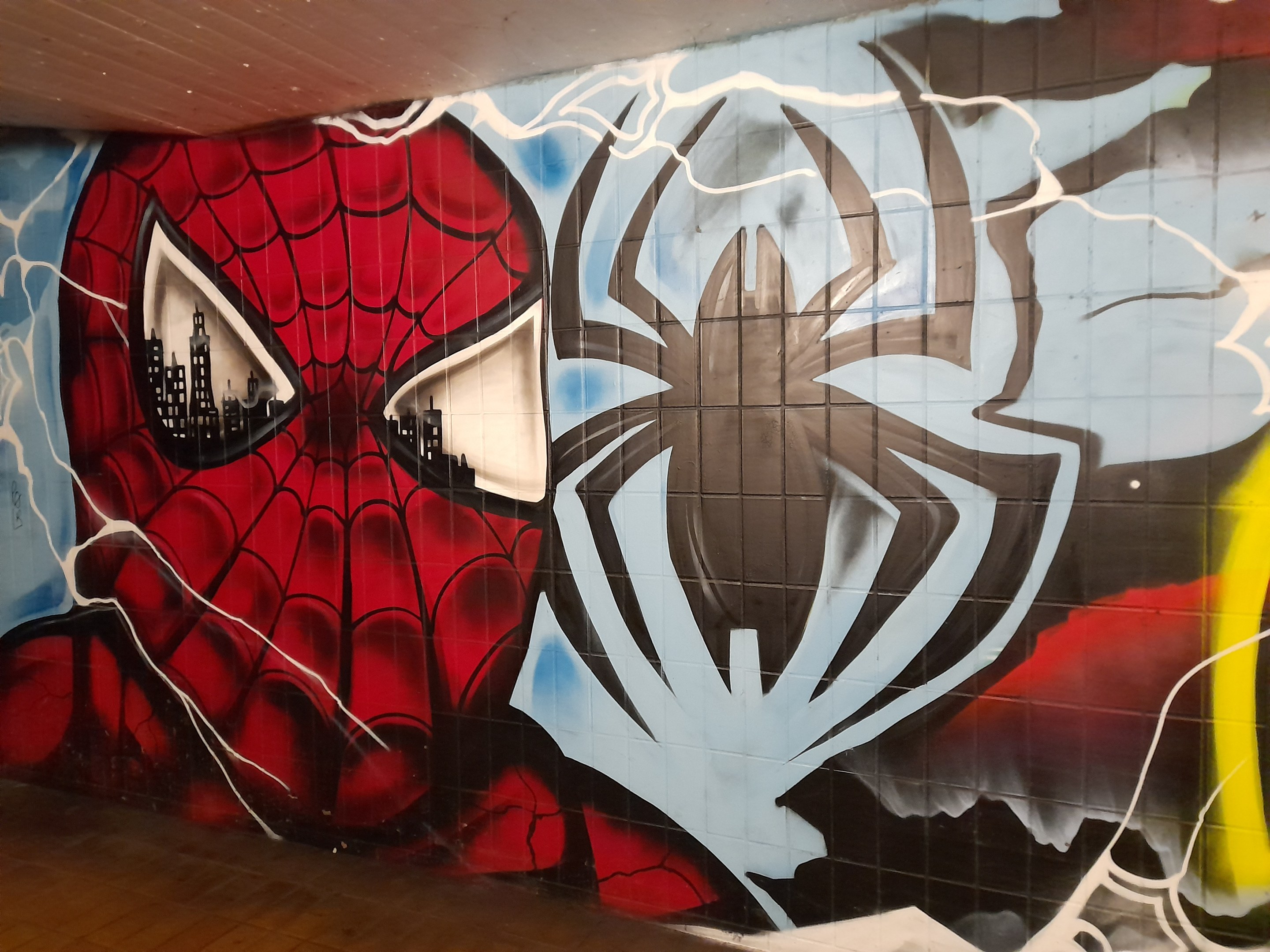 Mural von Spider-Man in rot und blauem Kostüm mit Webmuster und Spider-Logo auf einer Gebäudewand, mit breiteren Augen und leicht geöffnetem Mund.