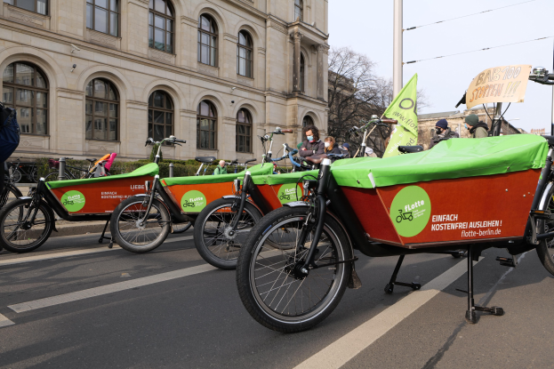 Eine Gruppe von Fahrrädern, die entlang einer Straße geparkt sind, mit einer Person in der Nähe und einem Banner, das eine Fahrrad-Sharing-Kampagne anzeigt, sowie Gebäuden, Bäumen und einem klaren Himmel im Hintergrund.