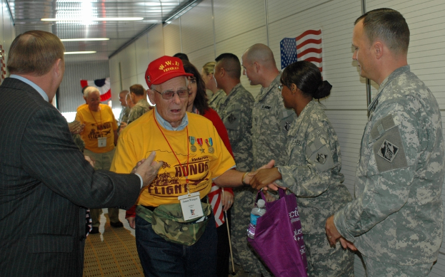 Eine Gruppe von Menschen und Soldaten steht drinnen zusammen, mit einer Flagge an der rechten Wand und Lampen an der Decke.