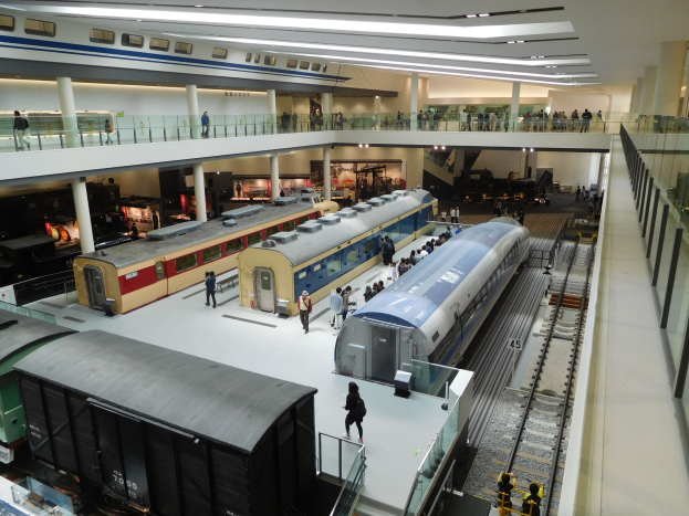 Eine große Museumsausstellung zeigt verschiedene Züge in unterschiedlichen Größen und Farben, mit Menschen, die von verschiedenen Ebenen und der Decke mit Beleuchtung beobachten.