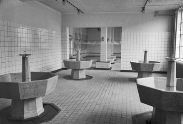 Schwarzes und weißes Foto einer öffentlichen Toilette mit mehreren Waschbecken auf Bodenhöhe, einer Wand im Hintergrund, Deckenleuchten und einem Fenster auf der rechten Seite.