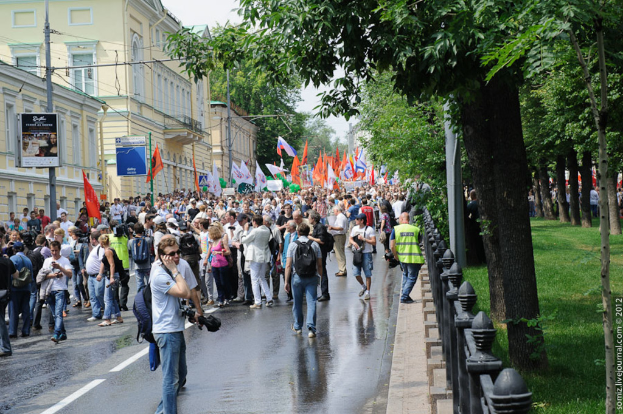 Eine Gruppe von Menschen steht auf einer Straße außerhalb der Stadt, viele halten weiße und orangefarbene Fahnen, mit verschiedenen Gebäuden und Bäumen im Hintergrund.