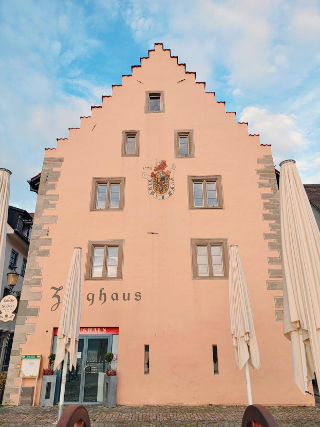 Ein pinkes Gebäude mit einem zentralen Uhrenturm, umgeben von Fenstern, Schirmen, einem Schild, einer Straßenlaterne, Topfpflanzen und einem bewölkten Himmel, repräsentiert München, Deutschland.