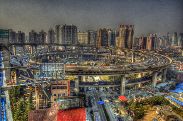 Stadtpanorama mit zentralem Ringstraßenverkehr, darunter Straße mit Autos und Fahrrädern, zahlreichen Hochhäusern im Hintergrund, Himmel oben, Bäumen links unten und verstreuten kleinen Häusern in der Stadt.