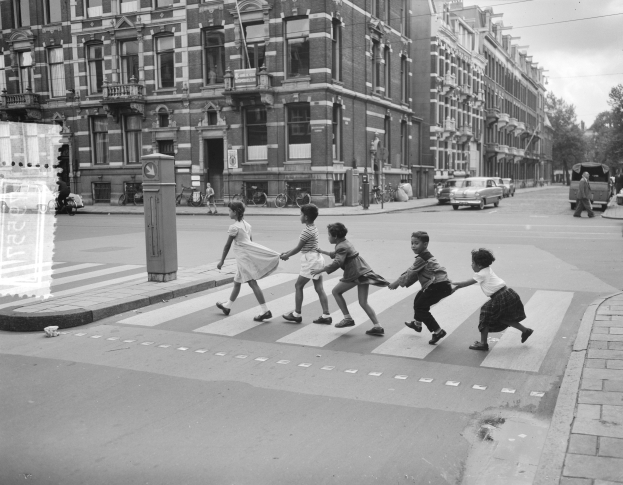 Eine Gruppe von Kindern überquert eine Straße vor einem Gebäude, mit Fahrzeugen, Fußgängern, Fahrrädern, Pfosten und Bäumen in einer schwarz-weißen Szene unter einem klaren Himmel.