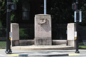 Ein Denkmal steht in der Mitte einer Stadtstraße, umgeben von Pfählen, Verkehrszeichen, Schildern, Geländern, Gras und Bäumen, mit einem Gebäude im Hintergrund, das Fenster aufweist.