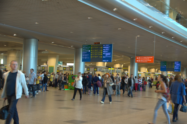 Menschen mit Gepäck durch den Flughafen gehen, umgeben von Informationsanzeigen, Deckenleuchten, Säulen und einem Glasgeländer.
