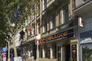 Eine belebte Stadtstraße mit einem hohen Gebäude in der Mitte, auf dem "CC Deluxe Cafe & Cocktail Bar" steht, umgeben von Fahrzeugen, Fußgängern, Gebäuden, Straßenmöbeln und einem klaren blauen Himmel.