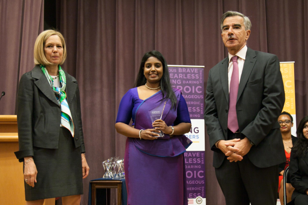 Zwei Frauen und ein Mann stehen auf einer Bühne mit einem braunen Vorhang dahinter; die Frau in der Mitte hält einen Award, und mehrere Awards sind auf einem Tisch vor ihnen.