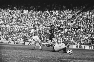 Schwarzes und weißes Foto von Männern, die auf einem Feld Fussball spielen, mit Zuschauern im Hintergrund und einer 'Bundesliga 1978-79 Ajax 1-1 Hannover 96'-Fahne.