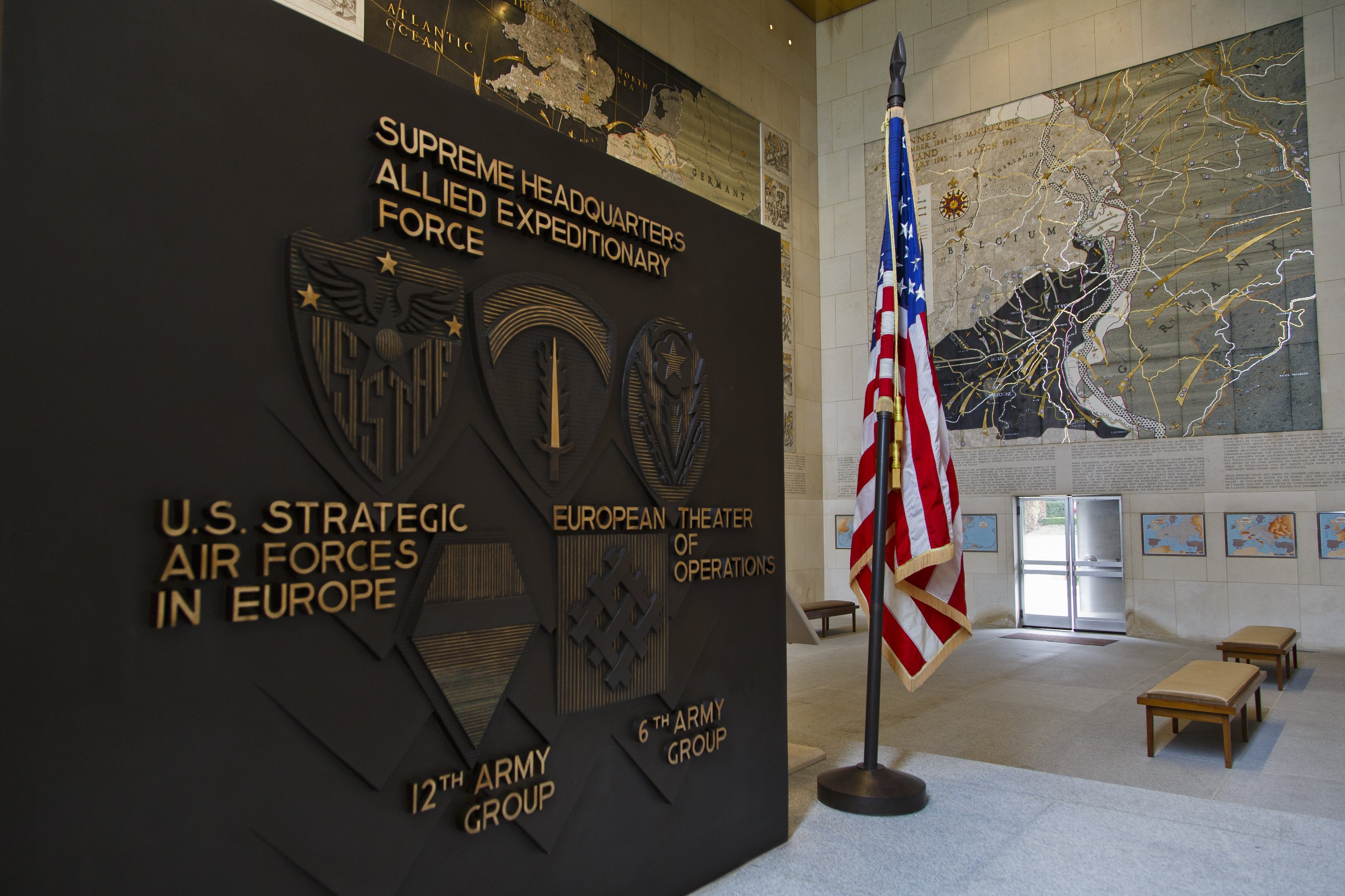 Eintritt zum U.S. Strategic Air Forces Headquarters in Europa mit einer amerikanischen Flagge, Text und Logos an der Wand, Bänken, einer Tafel, Fotorahmen, beleuchteter Decke und einer Tür im Hintergrund.