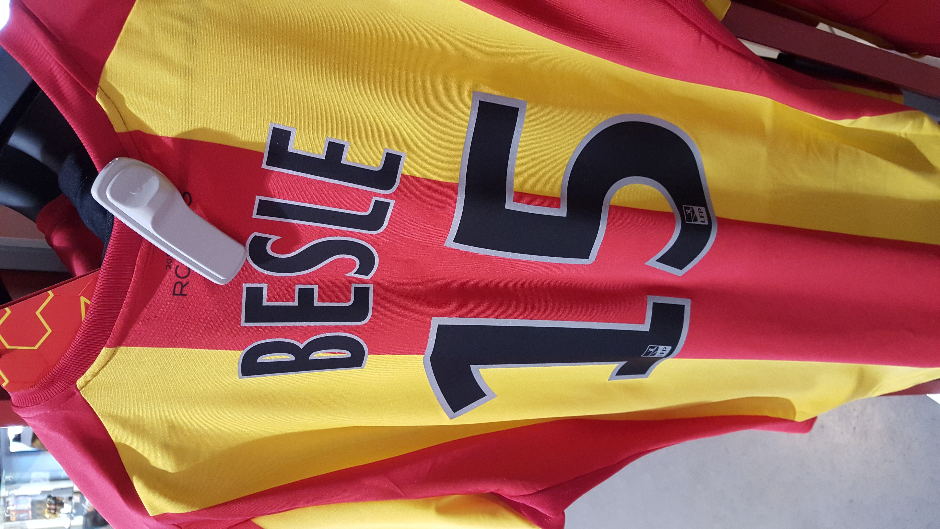 Rotes und gelbes Fußballtrikot mit aufgedrucktem Text und Nummern, das an einer Ladengestell hängend mit anderen Gegenständen und dem Hintergrund im Hintergrund zu sehen ist.