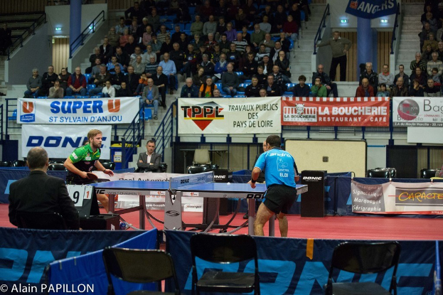 Indoor-Tischtennis-Match mit zwei Spielern, die gegeneinander antreten, sowie einigen stehenden Zuschauern, sitzenden Zuschauern, leeren Stühlen und Werbetafeln in einer Stadionatmosphäre.