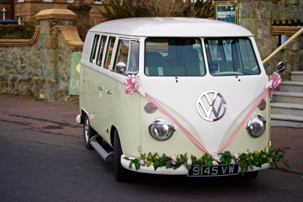 Ein weißer und pinkfarbener VW-Campervan mit floralen Verzierungen, der vor einer Steinmauer geparkt ist, neben einer Treppe und einem Gebäude im Hintergrund.