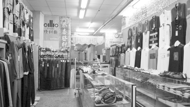 Eine Ausstellung von Kleidung, Gürteln und Schuhen mit einer Uhr an der Wand, hängendem Stoff im Hintergrund, Lampen darüber und einem Banner mit Text auf der linken Seite.