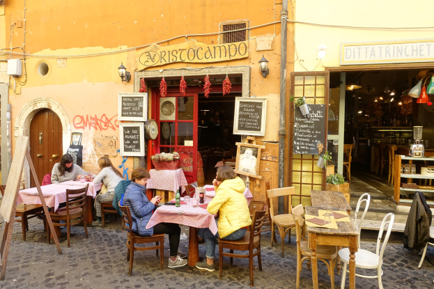 Eine Gruppe von Menschen sitzt an Tischen vor einem Restaurant in Rom, Italien, mit Flaschen, Gläsern und anderen Gegenständen auf den Tischen und Wänden mit Texttafeln, einer Tür ins Innere und beleuchteten Lichtern, sowie Regalen mit verschiedenen Gegenständen im Inneren.