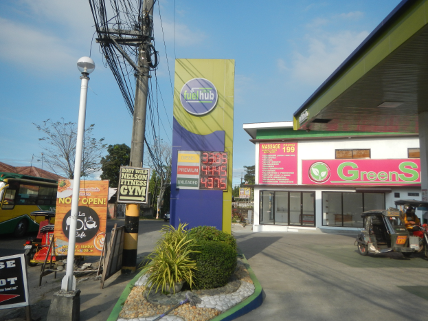 Eine Tankstelle mit dem Schild "Green's" davor, Fahrzeuge auf der Straße, verschiedene Strukturen und Vegetation im Vordergrund und Bäume und Wolken im Hintergrund.