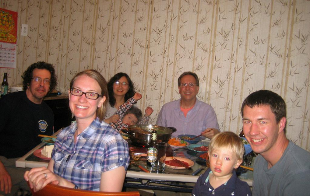 Eine Wohnzimmer-Szene mit einem Tisch in der Mitte, der mit Speisen gefüllt ist, zwei lächelnde Personen rechts, ein Mann und eine Frau links, drei Personen im Hintergrund mit einer Frau, die ein Kind hält, einem Kalender und einer Flasche oben links, floralen Wanddesigns und einem Stuhl im Vordergrund.