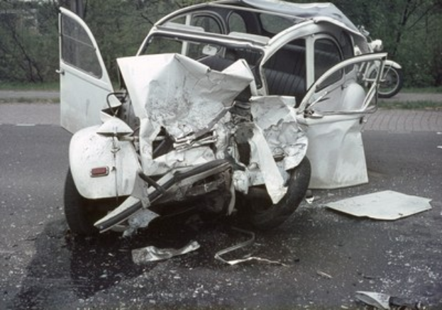 Stark beschädigte Limousine am Straßrand mit Frontalcrash, Bäume und ein Gebäude im Hintergrund, weiteres Fahrzeug in der Ferne sichtbar.