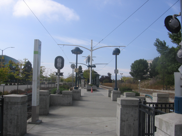 Ein Fußgängerbrücke mit Geländern, Pfählen und Laternen, umgeben von Pflanzen, Bäumen und Gebäuden, mit einem Zug auf den Schienen in der Nähe unter einem bewölkten Himmel.