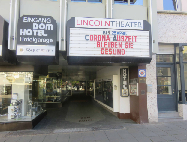 Das Lincoln Theater in Berlin, Deutschland, ein Gebäude mit Glasfenstern und -türen und einer Tafel mit Text darauf, mit verschiedenen Objekten im Inneren, die den Eindruck einer pulsierenden Stadtlandschaft vermitteln.