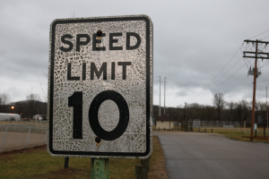 Geschwindigkeitsbegrenzungsschild am Straßenrand mit umgebenem Gras, Strommasten, einem Zaun, Häusern, Bäumen und einem bewölkten Himmel im Hintergrund.