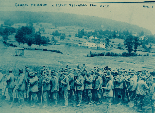 Eine Gruppe deutscher Kriegsgefangener in Frankreich mit Waffen in der Hand auf dem Boden stehend, umgeben von Bäumen, Häusern und einem klaren blauen Himmel.
