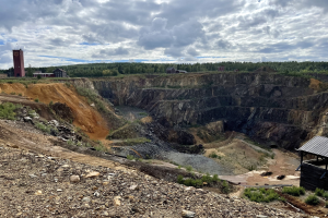 Großer Tagebau in einem Schmutzfeld umgeben von Bäumen, Gebäuden und einem Schuppen unter einem bewölkten Himmel mit verstreuten Steinen und Pflanzen.