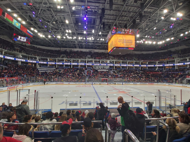Eine große Menschenmenge in einer Hockeyarena, einige sitzen und einige stehen, mit einem Zaun um die Eisfläche und Bannern, die von der Decke hängen, unter hellen Lichtern.