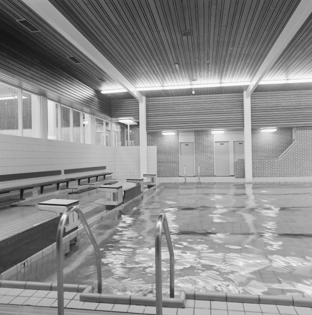 Große Hallenbad mit klarem Wasser, gefliestem Boden, Metallstangen, Bönchen, Wönden, Türen, Lampen und Fenstern, die die Decke zeigen.