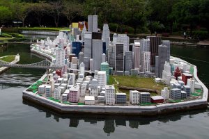 Ein Miniaturstadtmodell auf Wasser mit Gebäuden, Grünflächen, einer Brücke und Menschen, mit einem Himmelhintergrund.