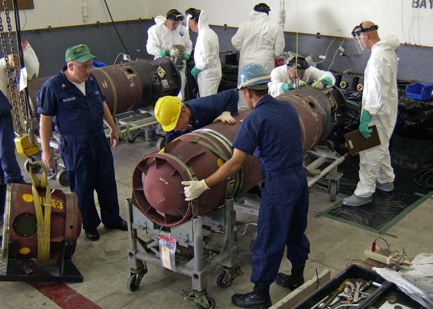Mannergruppe in Helmen und Handschuhen beim Entschärfen einer großen Bombe in einer Fabrik mit Maschinen und einer textbedeckten Wand im Hintergrund.