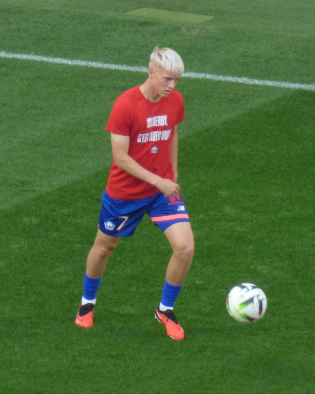Ein junger Mann in rotem Trikot und blauen Shorts, der auf einem grünen Rasen Fußball spielt, trägt Fußballschuhe und -bekleidung während eines Spiels.