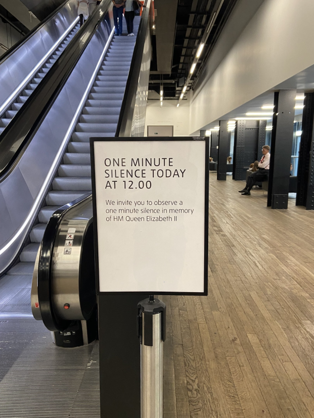 Eine Rolltreppe in einem Flughafen mit einem Schild, auf dem "Eine Minute Stille heute" steht, einige Menschen darauf und an der Decke angebrachte Lampen im Hintergrund.