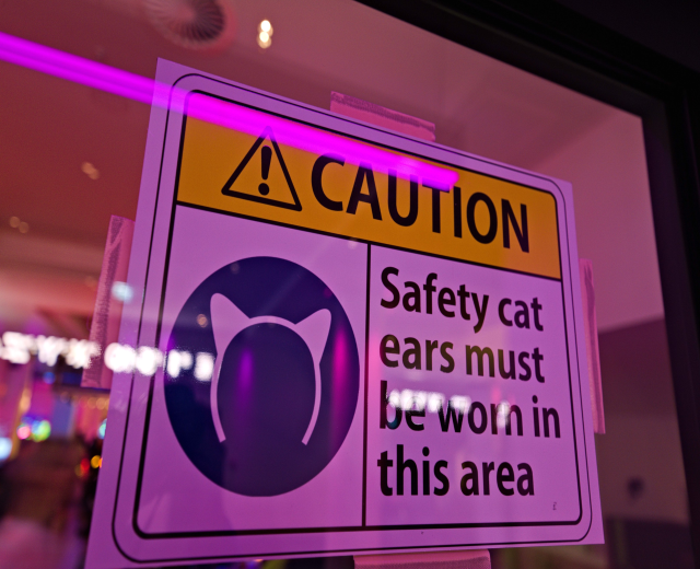 Hellgelbes Warnschild in einem Ladenschaufenster mit schwarzem Text, der "Vorsicht: Sicherheitskatzenohren müssen in diesem Bereich getragen werden" lautet.