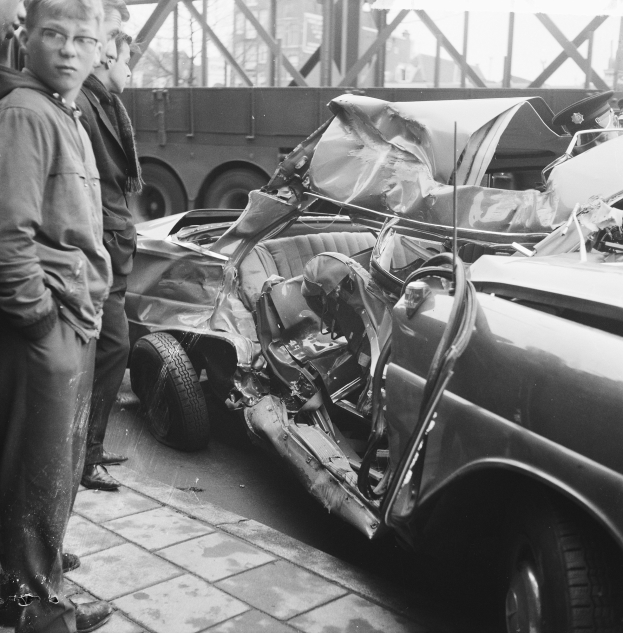 Ein Schwarz-Weiß-Bild eines verunglückten Autos am Straßenrand mit mehreren Menschen drumherum, einem Lastwagen und einer Brücke im Hintergrund.