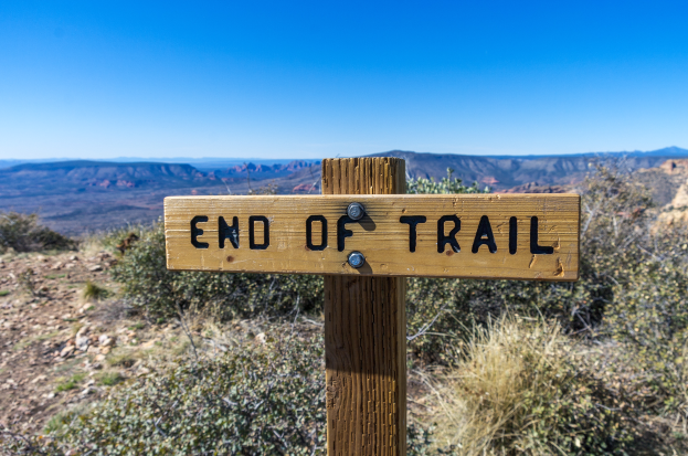 Ein hölzernes Schild mit der Aufschrift "Ende des Weges" ist an einem hölzernen Pfahl im Vordergrund befestigt, umgeben von Pflanzen und Hügeln im Hintergrund unter einem klaren blauen Himmel.