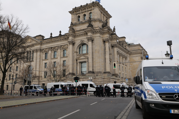 Eine Gruppe von Polizisten steht vor dem Reichstaggebäude in Berlin, Deutschland, mit Fahrzeugen, einem Zaun, Verkehrsampeln, Laternen, Bäumen und Fahnen im Hintergrund, bei klarem Himmel.