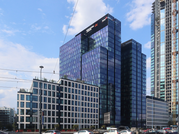 Großes modernes Gebäude mit vielen Fenstern, als Deutsche Bank Hauptsitz in Frankfurt, Deutschland identifiziert, umgeben von städtischer Infrastruktur einschließlich Straßenlaternen, Fahrzeugen, Bäumen und einem bewölkten Himmel.