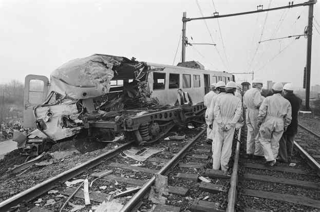 Ein Schwarz-Weiß-Bild einer Zugkollision mit Menschen in Helmen, die herumstehen, Strommasten, Bäumen, Gebäuden und einem klaren Himmel im Hintergrund.