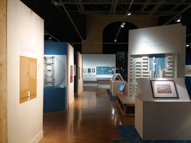 Ein Raum mit verschiedenen Kunstwerken, darunter gerahmte Fotos auf Tischen, Texttafeln an Wänden, Deckenleuchten und ein Teppichboden, typisch für eine Ausstellung im Museum of Modern Art.