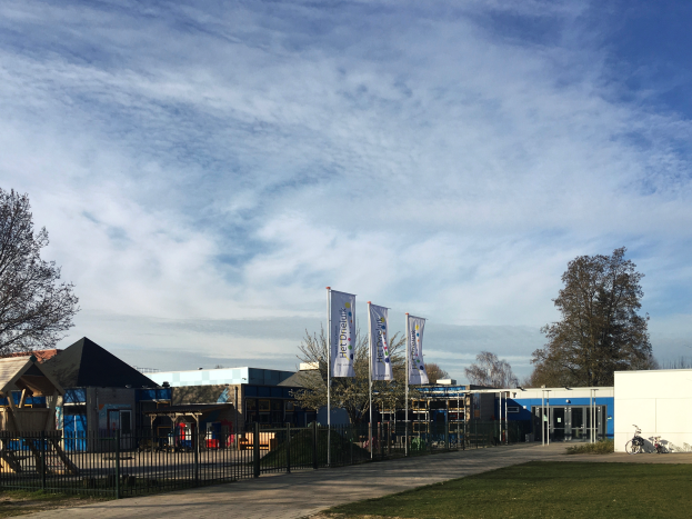 Ein großes Gebäude mit einer grasbewachsenen Fläche davor, umgeben von einem Metallzaun, einem Weg, Bäumen, Pfosten, Bannern, einem geparkten Fahrrad, nahen Häusern und einem bewölkten Himmel, das als neuer Spielplatz an einer Schule dient.
