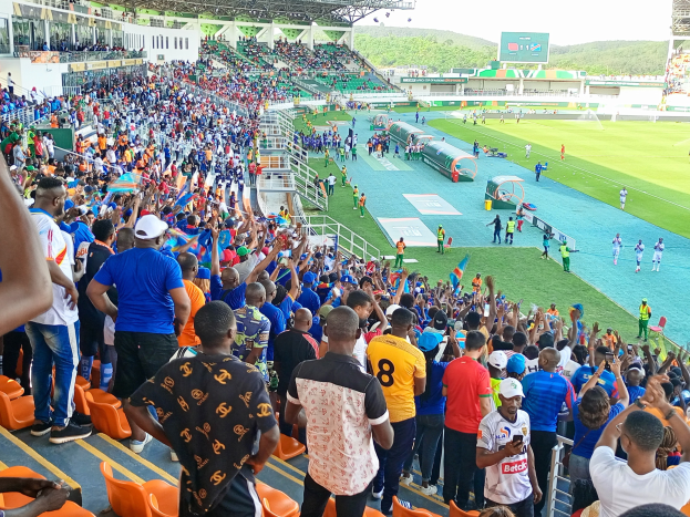 Eine große Menschenmenge versammelt sich vor einem eingezäunten Fußballfeld, mit einigen auf Stühlen sitzend und anderen stehend, mit Blick auf Hügel und einen klaren blauen Himmel während der 2019 African Cup of Nations.