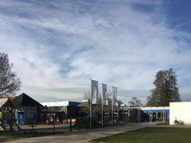 Großes Gebäude mit einem Metallzaun, Rasenfläche, Weg, Bäumen, Schildern, einem Fahrrad, nahen Häusern und einem bewölkten Himmel, identifiziert als der neue Spielplatz an der Schule.