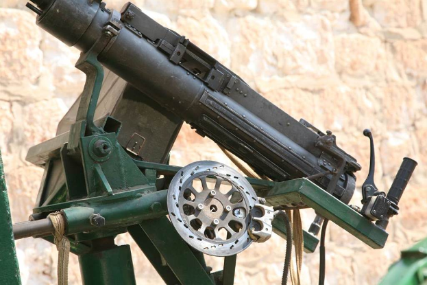 Eine Maschinengewehr auf einem Stahlständer mit einem Griff auf der rechten Seite, Seile und Kabel unten und eine Wand im Hintergrund.