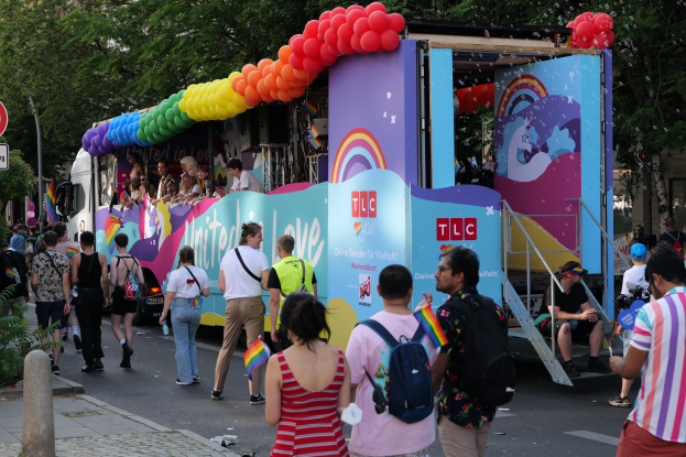 Eine Gruppe von Menschen, die eine Straße entlanggehen, neben einem Lastwagen mit bunten Luftballons, mit Schildern an der Straße und Bäumen und Gebäuden im Hintergrund, was auf eine Pride-Parade in Paris hindeutet.