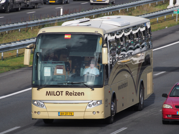 Ein Minibus mit Passagieren auf einer Autobahn neben einem roten Auto, mit Leitplanken und Gras am Straßenrand und Texttafeln im Hintergrund.