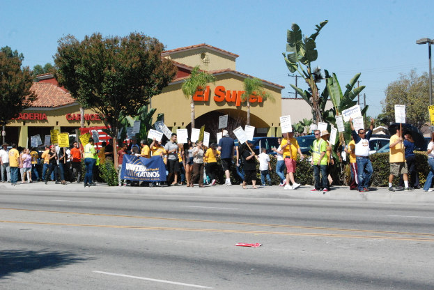 Eine Gruppe von Menschen mit Schildern, die durch einen Park gehen, mit Bäumen, Büschen, einer Fahne, einem Gebäude mit Namensschildern, Autos und aktuellen Umfragen im Hintergrund.