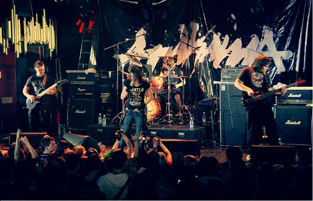 Eine Gruppe von Musikern, die auf der Bühne mit Gitarren und Trommeln spielen, Mikrofone und Ständer sowie ein Banner im Hintergrund und Text am unteren Rand, der "The Wanted - Live at the O2 Academy, London" lautet.