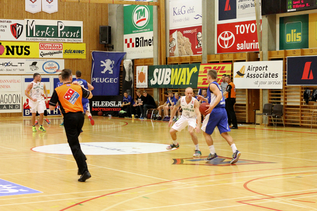 Ein Basketballfeld mit Spielern im Vordergrund und einer holzgetäfelten Wand mit Postern und Werbetafeln im Hintergrund.